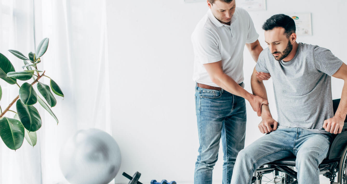 A caregiver assists a young man attempting to stand up from his wheelchair